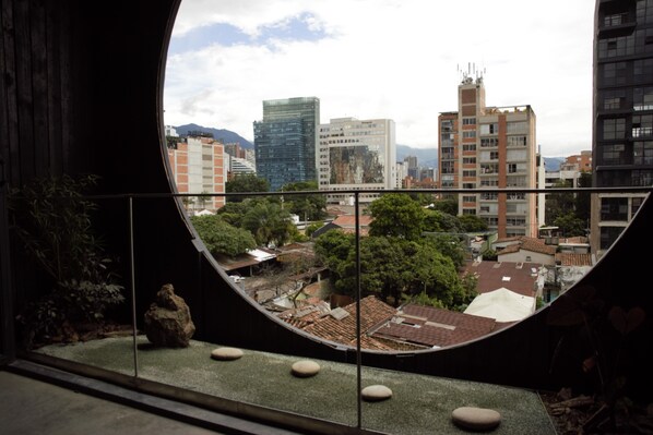 A view through a circular window of a cityscape with modern buildings, a rooftop garden, and a large rock centerpiece.