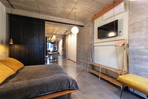 Une chambre d’hôtel moderne avec un grand lit, un banc en bois, un téléviseur fixé au mur et un plafond en béton.