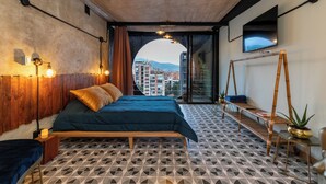 Un dormitorio con cama, televisor, ventana con vista a edificios y piso con diseño.