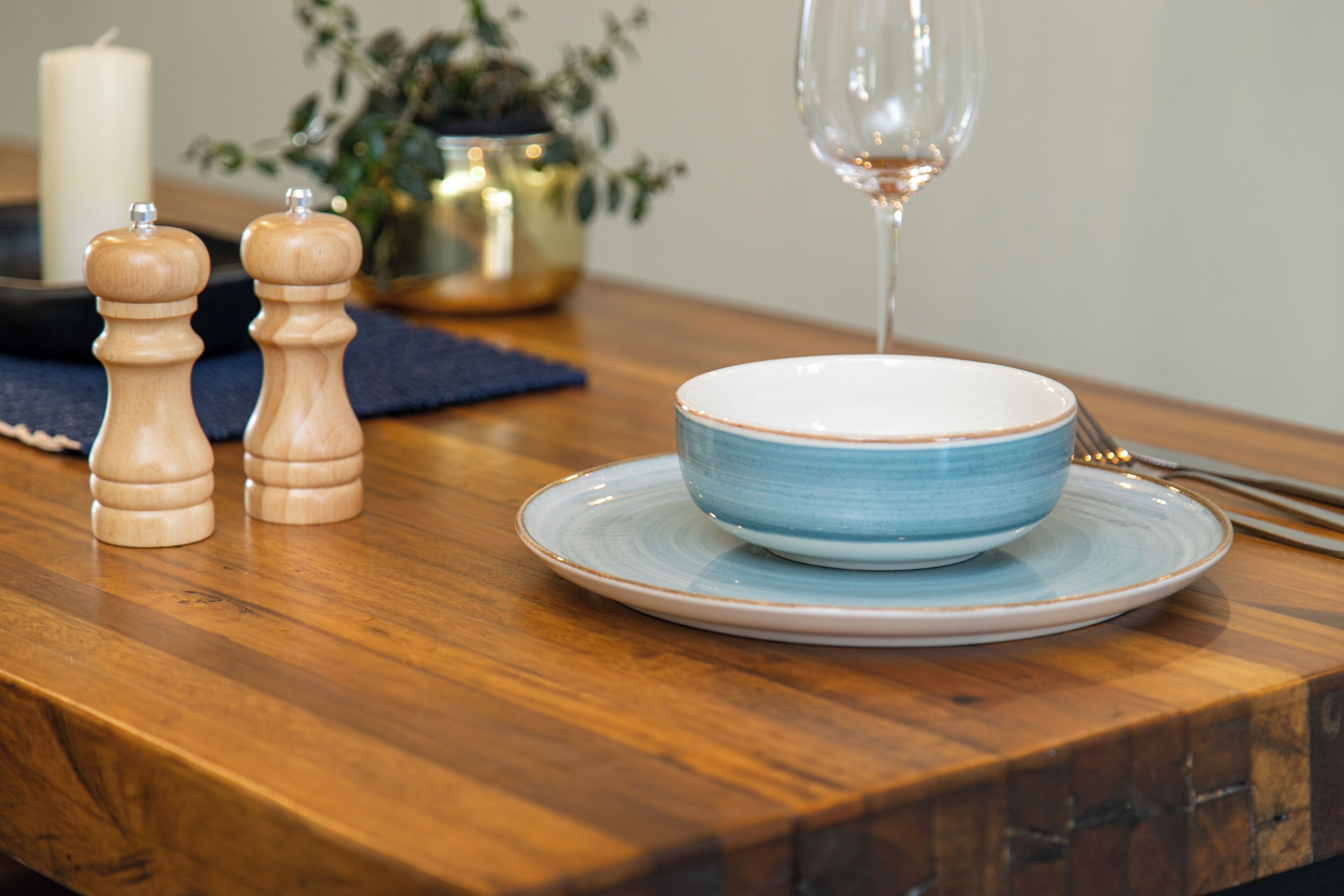Una mesa de madera con un cuenco azul y un plato, dos saleros y pimenteros de madera, una vela y una copa de vino.