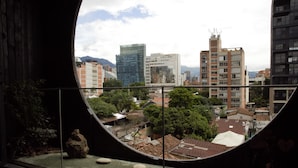 Vista a través de una ventana circular de un paisaje urbano con edificios modernos, un jardín en la azotea y una gran roca como pieza central.
