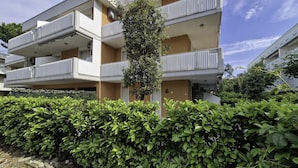 Un edificio residencial de varios pisos con balcones, un árbol central y un seto bien cuidado al frente.