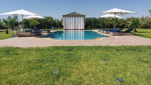 A swimming pool with lounge chairs and umbrellas, surrounded by a grassy area and a covered structure. - Saphir Paon - Luxury Villa in the Middle of a Farm (Souihla)