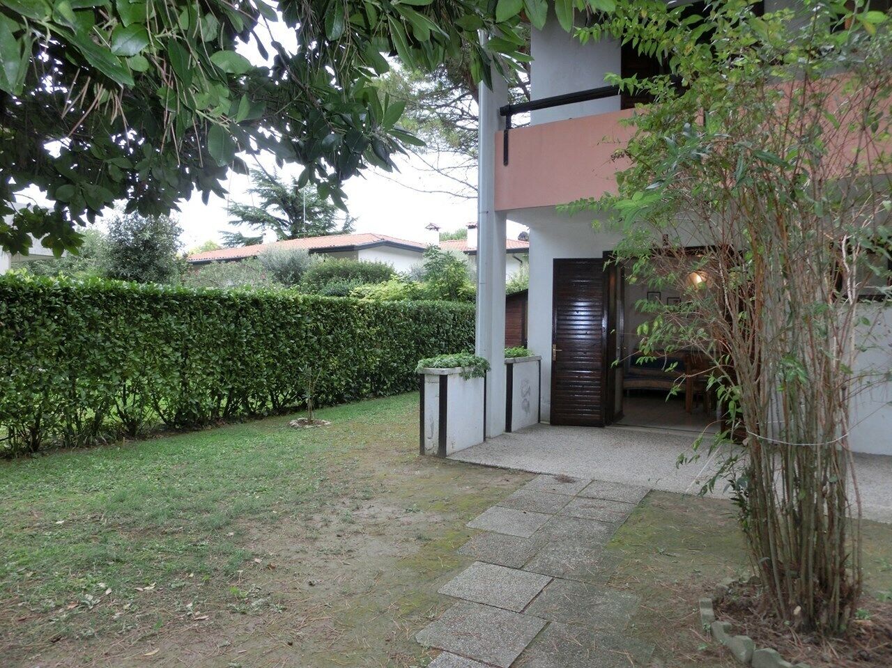 A modern building with a dark entrance door, a paved pathway leading to the entrance, and a well-maintained hedge in the background.