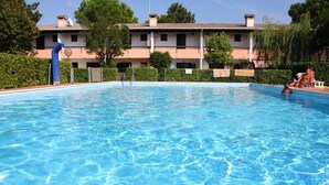 A rectangular swimming pool with clear blue water, surrounded by a low hedge and a building with a tiled roof in the background. - Two-bedroom Apartment With Private Garden (San Michele al Tagliamento)