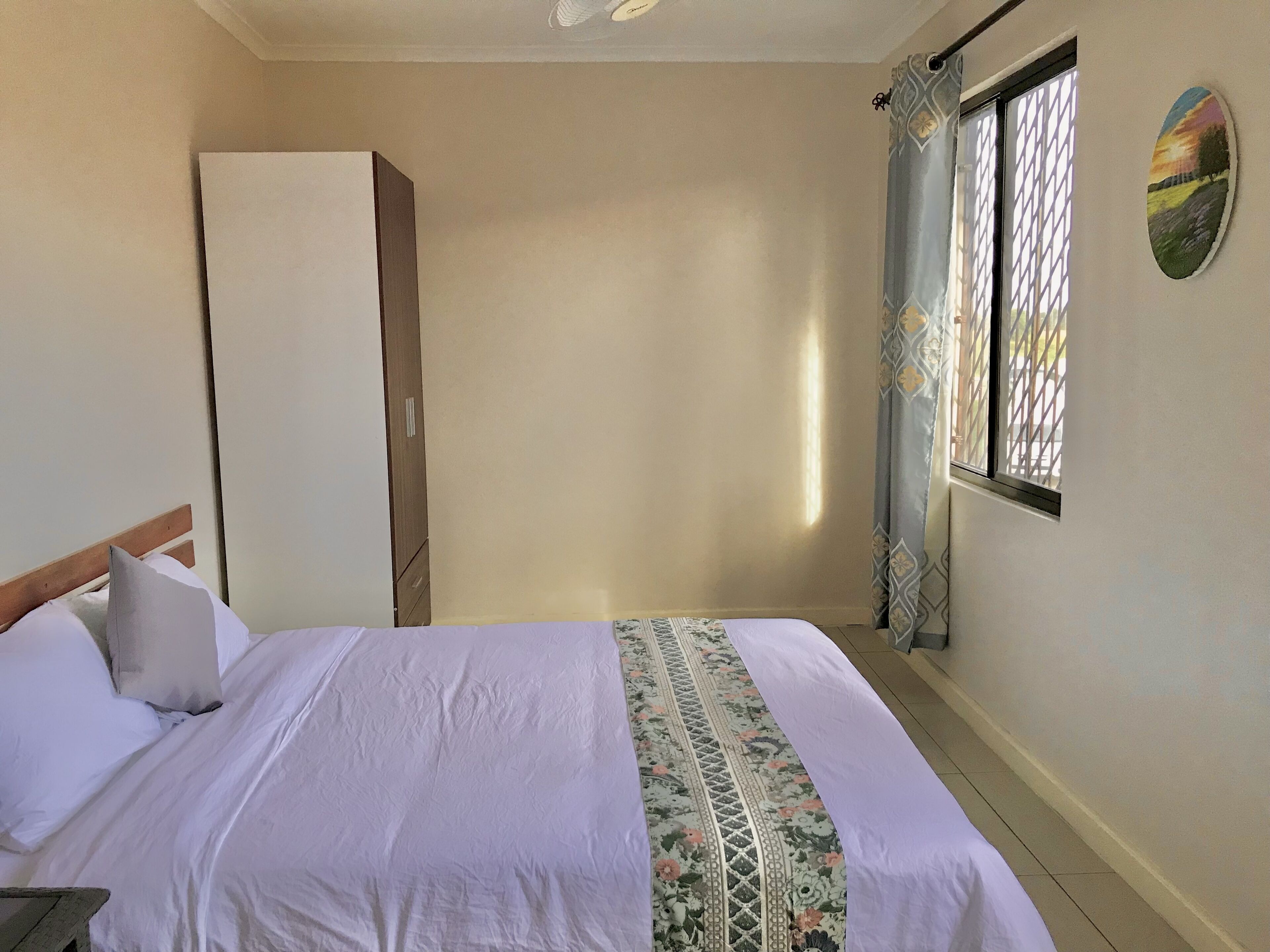 A bedroom with a bed, a wardrobe, a window with curtains, and a wall with a painting.