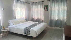 Une chambre à coucher avec un lit, des rideaux, une petite table sur laquelle se trouve un vase, et un tableau accroché au mur.