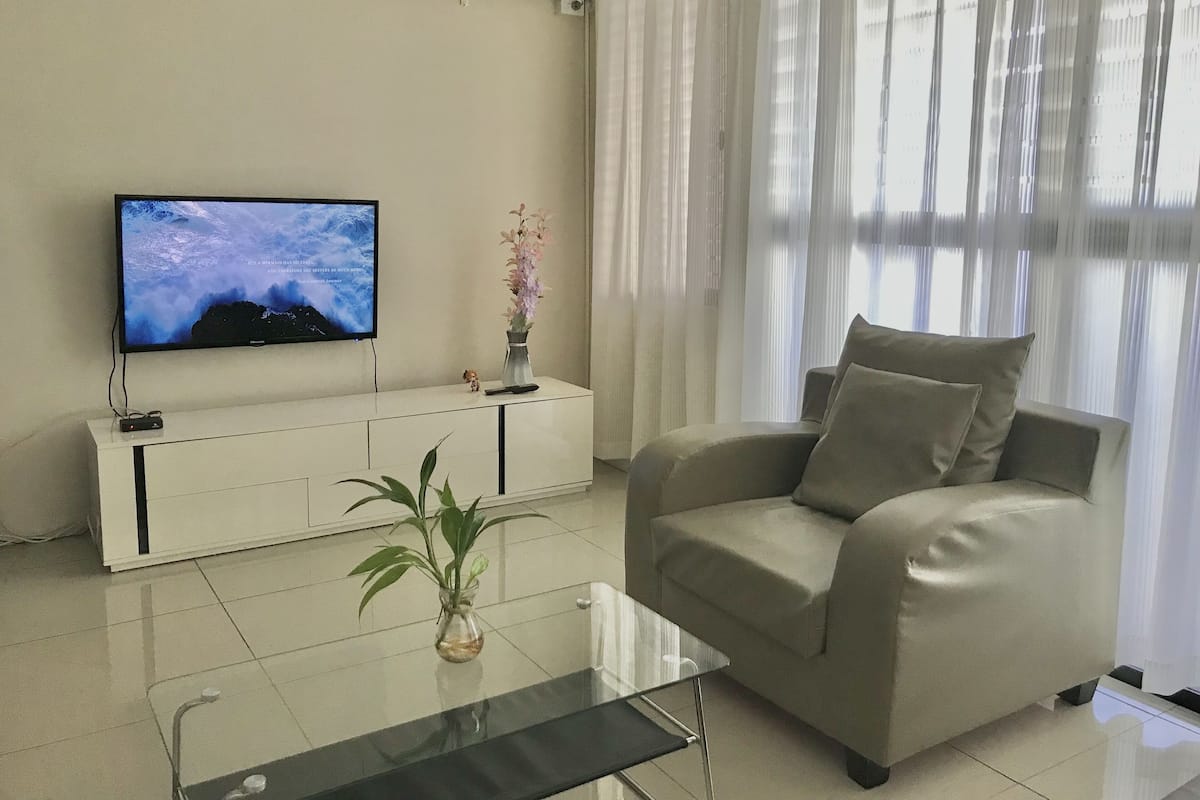 Una sala de estar con un sofá beige, una mesa de centro de vidrio, un televisor sobre un mueble blanco, un jarrón con flores y cortinas de gasa.