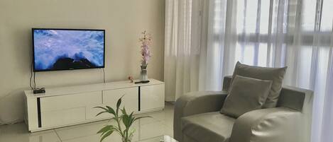 A living room with a beige sofa, a glass coffee table, a TV on a white cabinet, a vase with flowers, and sheer curtains.