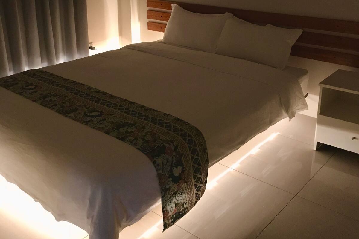 Una cama bien hecha con una colcha estampada, una mesita de noche y un cuadro en la pared.
