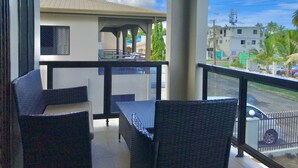 Un balcón con sillas de mimbre y una mesa, con vista a una calle con edificios y palmeras.