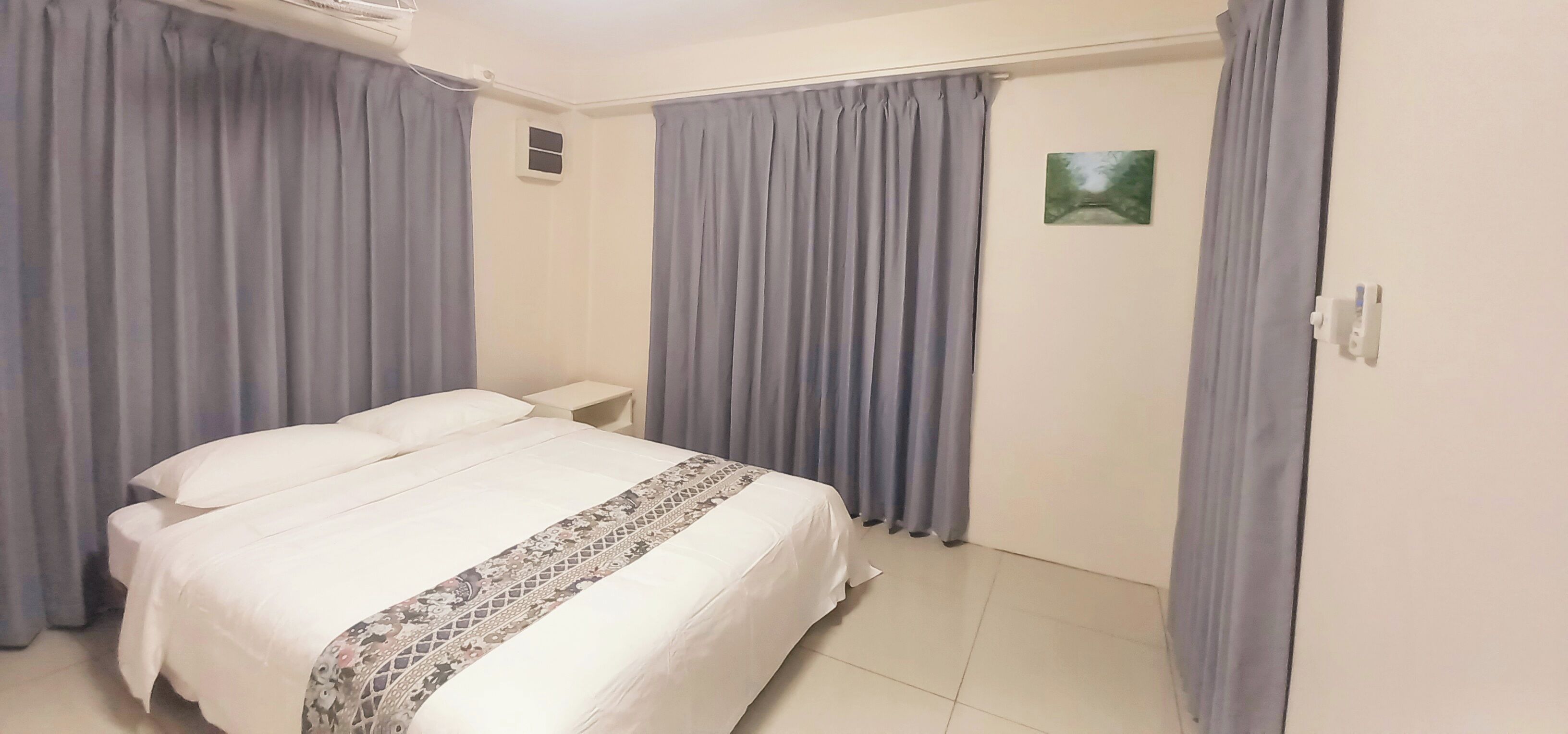 Ein Hotelzimmer mit Bett, Vorhängen, Nachttisch, kleinem Fenster und einem an der Wand befestigten Lichtschalter.