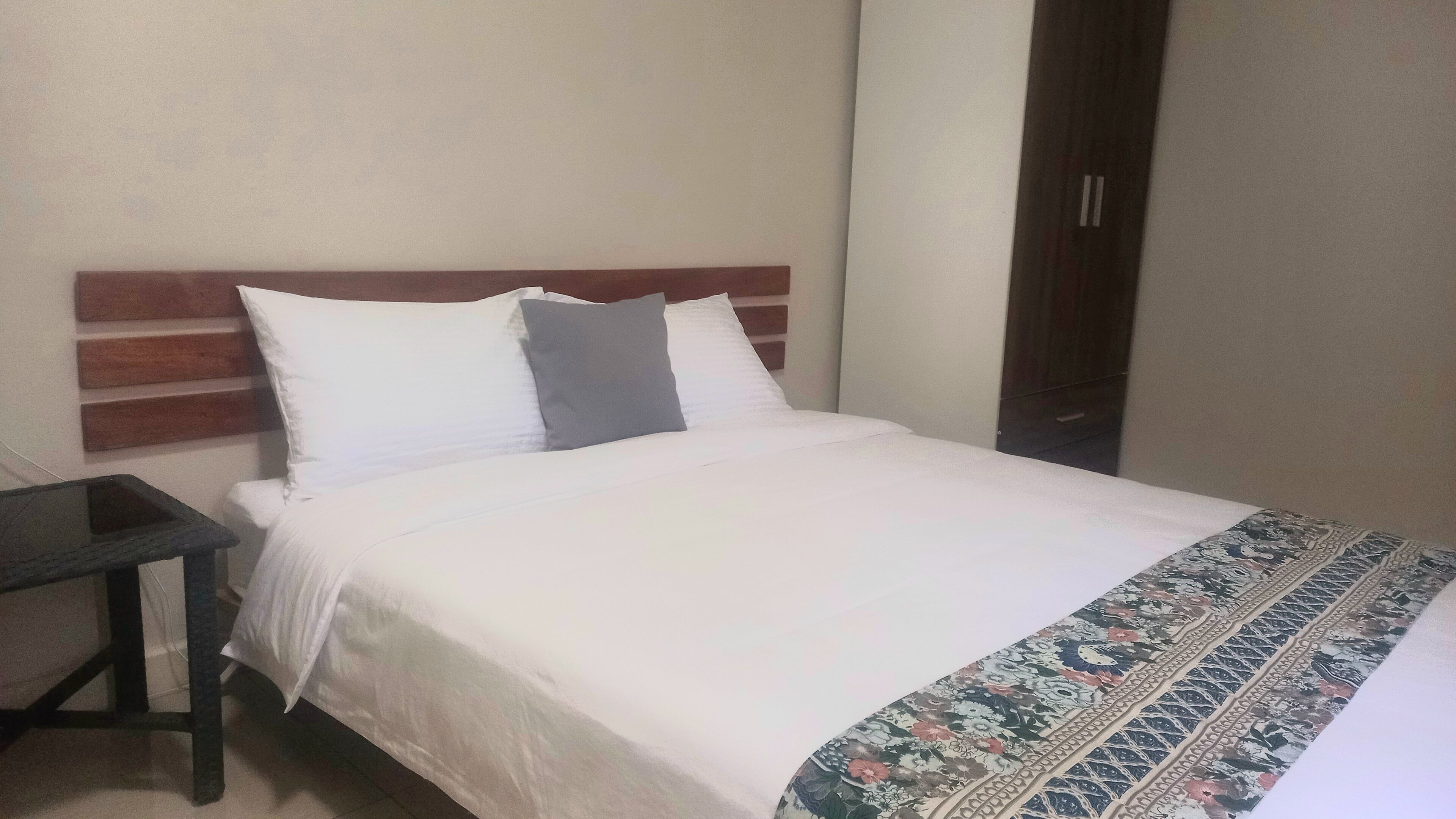 Una cama bien hecha con ropa blanca y una manta estampada. A la izquierda de la cama, hay una mesita. La habitación tiene un diseño sencillo y limpio, con un cabecero de madera y una pared lisa.