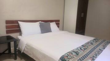 A neatly made bed with white bedding and a patterned throw. A small table is positioned to the left of the bed. The room has a simple, clean design with a wooden headboard and a plain wall.