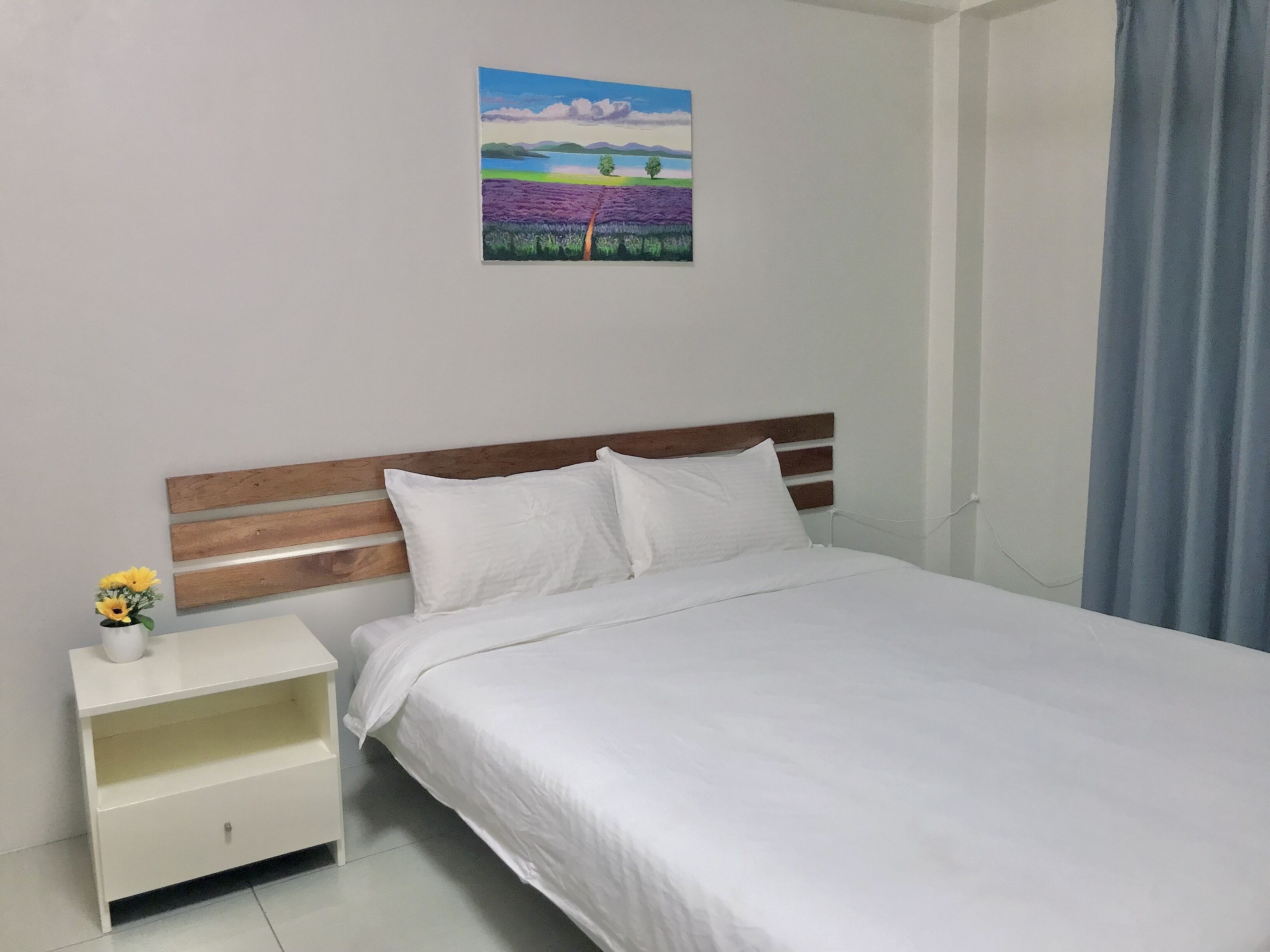 Una cama bien hecha con ropa blanca, un cabecero de madera y una mesita de noche con un florero de girasoles.