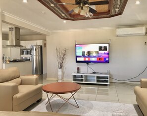Un salon moderne avec un téléviseur à écran plat, des canapés de couleur beige, une table basse ronde et un vase décoratif.