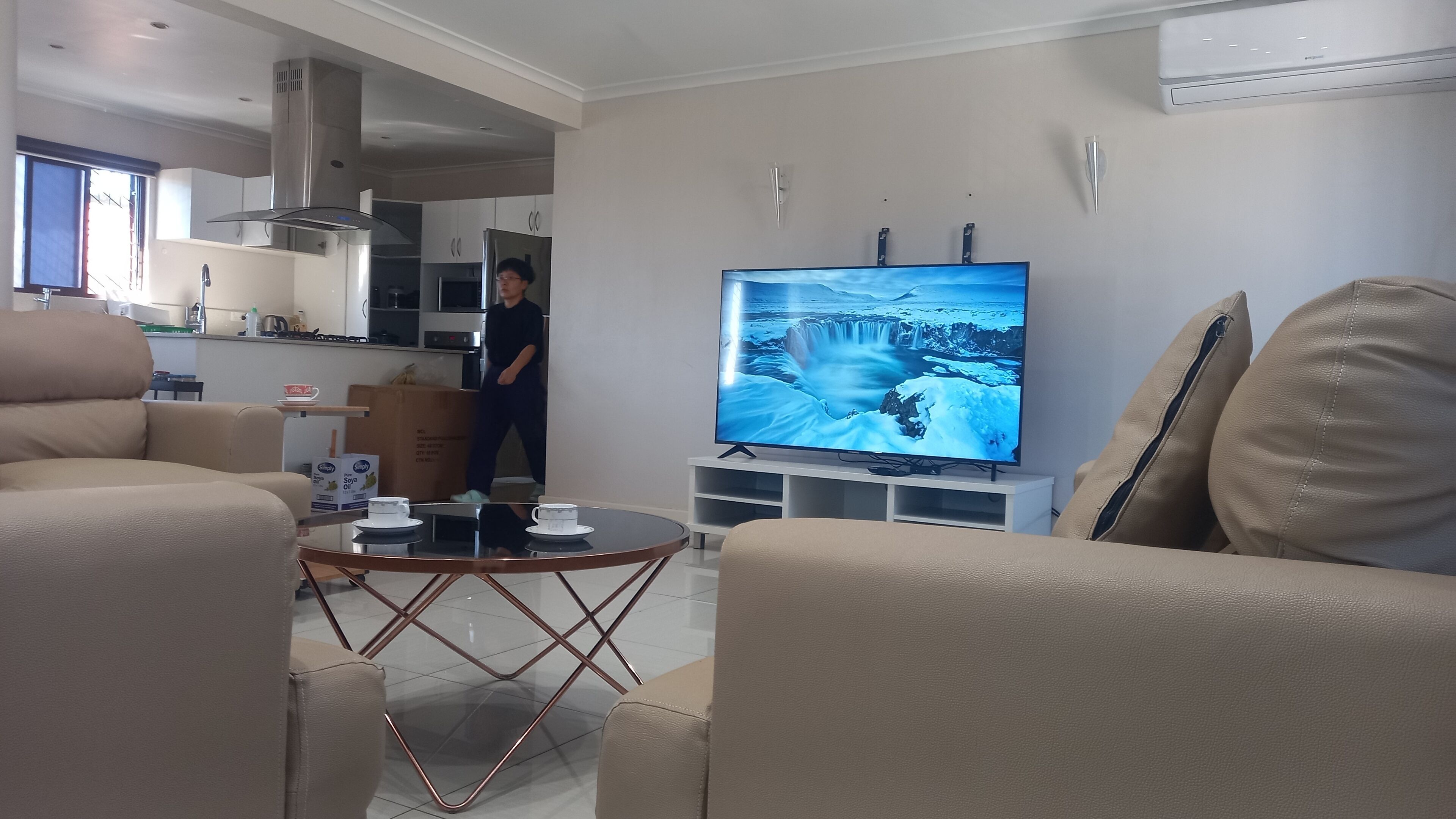 A modern living room with a large flat-screen TV, beige sofas, a glass coffee table, and a kitchen in the background.