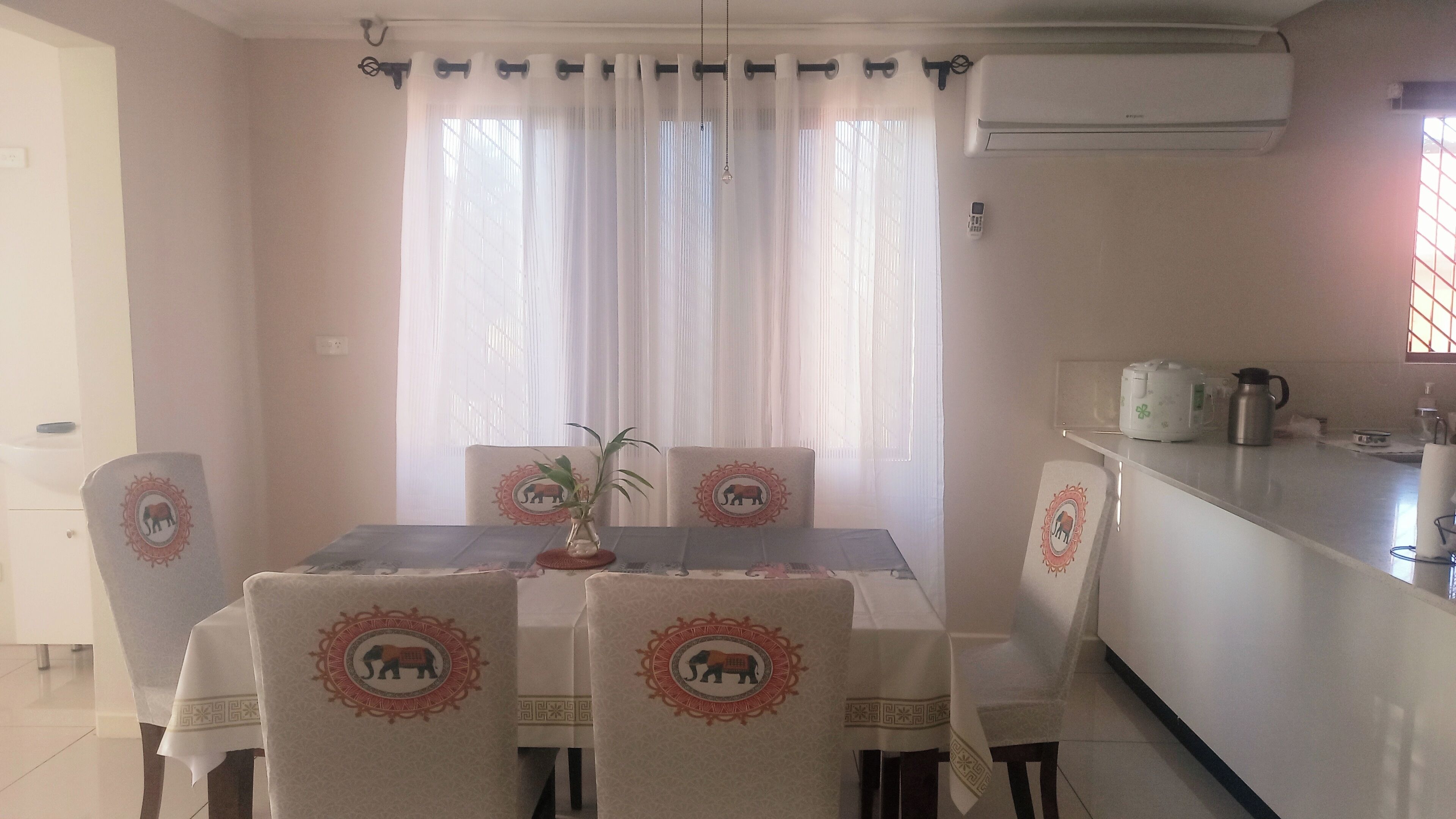 A dining area with a table set for four, chairs with elephant designs, a potted plant, and a kitchen counter with a kettle.