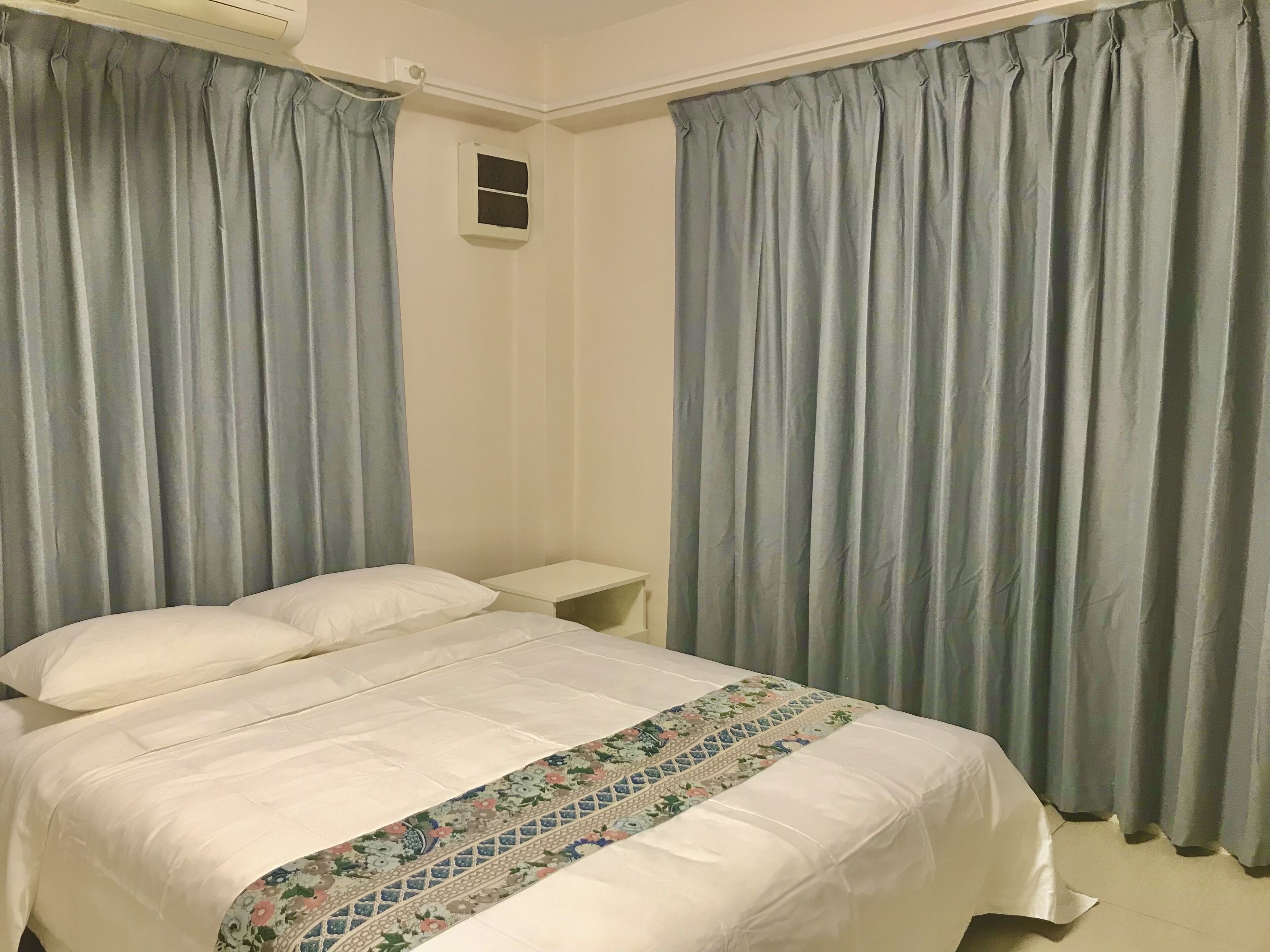 A bedroom with a bed, curtains, a nightstand, and a wall-mounted air conditioning unit.