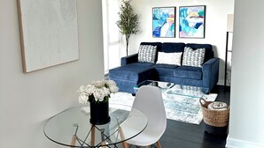 A modern living room with a glass dining table, white chairs, a blue sofa, and abstract art on the walls.