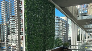 A balcony with a wooden bench and two chairs, overlooking a cityscape with modern buildings and a green wall.