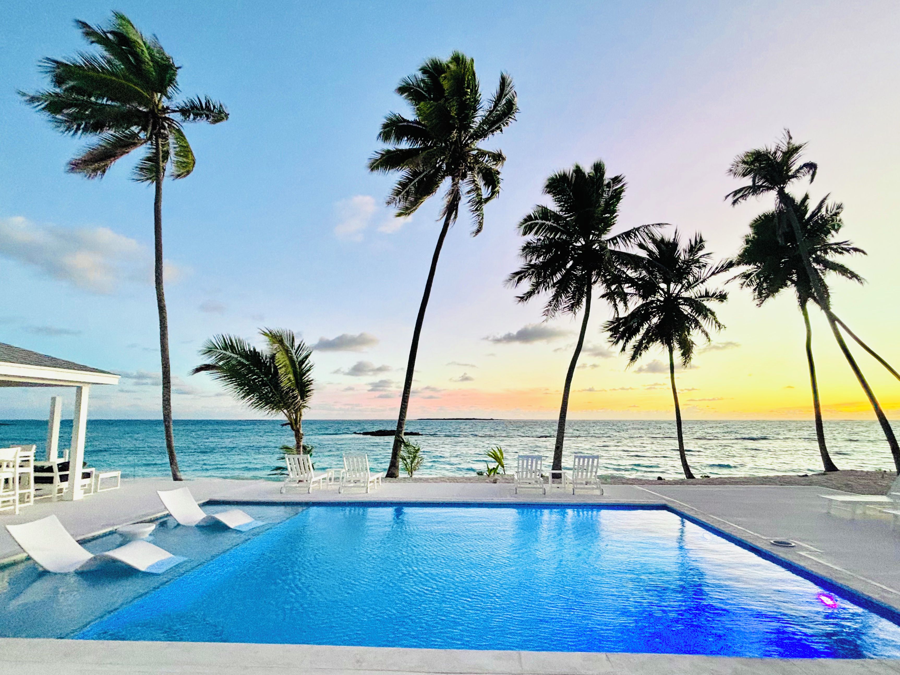 A swimming pool with lounge chairs, palm trees, and a view of the ocean at sunset.