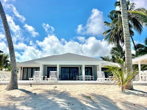 A beachfront property with a covered patio, white lounge chairs, and a sandy beach.