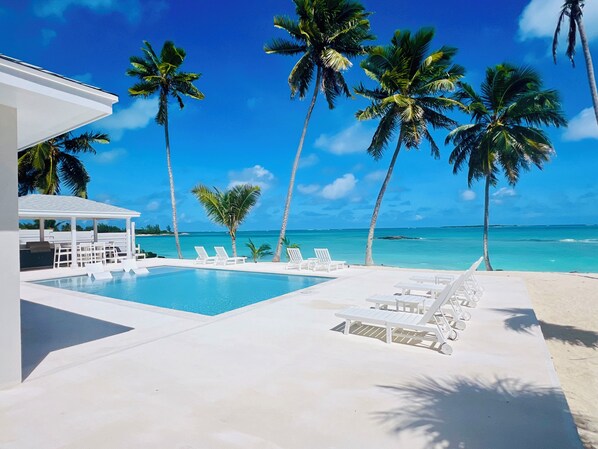 Un espace piscine avec des chaises longues, un coin salon abrité et des palmiers.
