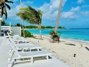 Un espace de détente aménagé sur la plage, avec des chaises longues et des tables blanches, des palmiers et une vue sur l’océan.