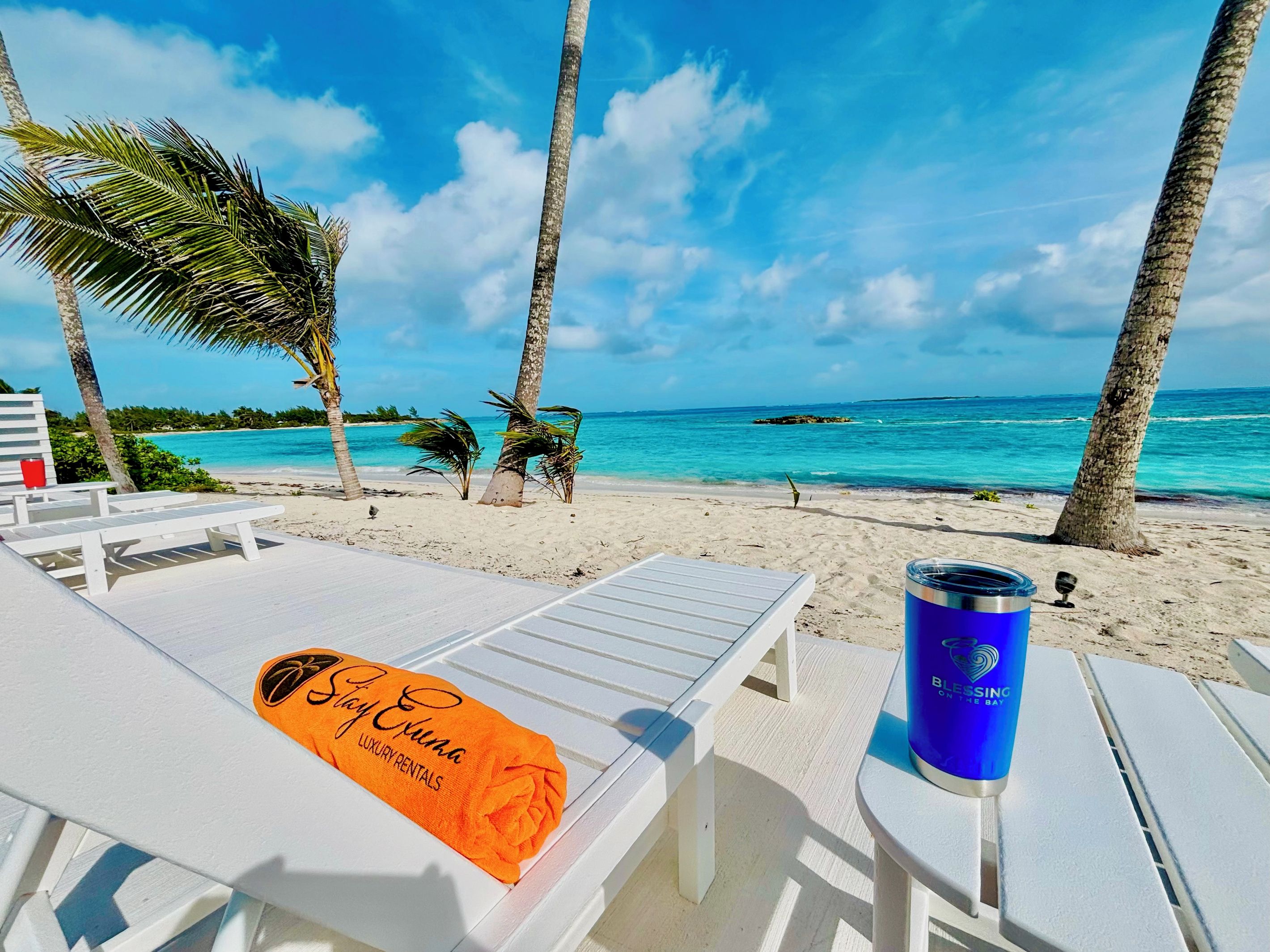 A beachside view with lounge chairs, a blue tumbler, and a towel with 'Stay Exuma Luxury Rentals' text.