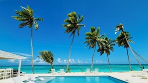 A pool area with white lounge chairs and a white pergola, surrounded by palm trees and a clear blue sky.