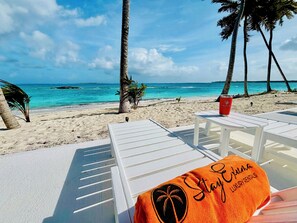 Un salon de plage avec des chaises longues blanches, un verre rouge et une serviette orange sur laquelle est inscrit le texte « Stay Puma Luxury Rentals ».