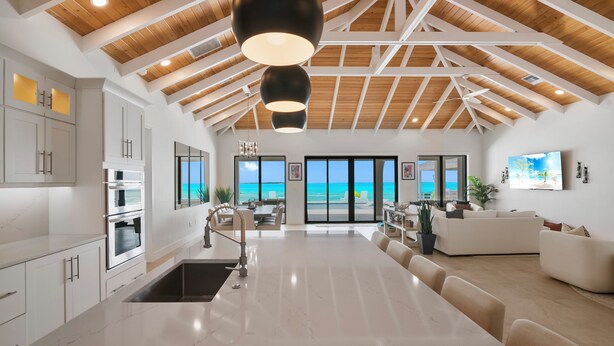 A modern kitchen with white cabinets and a large island, a dining area with a long table, and a living room with a white sofa and a TV.