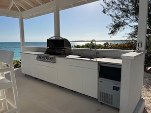 An outdoor kitchen with a grill, sink, and built-in refrigerator, overlooking a body of water. - Island Time - Brand New 4 bedroom Luxury House right on the Beach! (Steventon)
