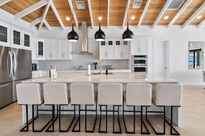 A modern kitchen with a central island, four bar stools, stainless steel appliances, and a wooden ceiling. - Island Time - Brand New 4 bedroom Luxury House right on the Beach! (Steventon)