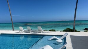 A pool with white lounge chairs and palm trees, overlooking a clear blue sea.