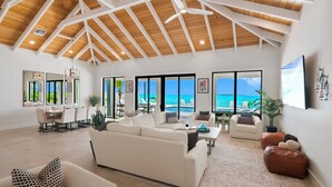 A spacious living room with a high ceiling, large windows offering ocean views, and a dining area with a view of the pool.