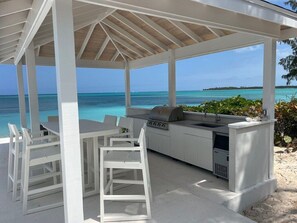 Cocina exterior con parrilla y lavamanos, mobiliario blanco y vista al océano.