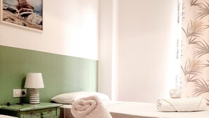 Una habitación de hotel con cama, una mesita de noche con lámpara, un cuadro de un barco en la pared y una ventana con cortina.
