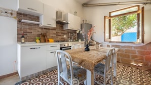 A compact kitchen with white cabinets, a brick backsplash, and a dining area with a patterned tablecloth and metal chairs. - La Casa sul Mare (Su Pallosu)