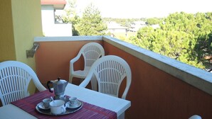 A balcony with a table set for two, white chairs, and a view of trees and buildings in the background. - Two-bedroom Apartment in Bibione (San Michele al Tagliamento)