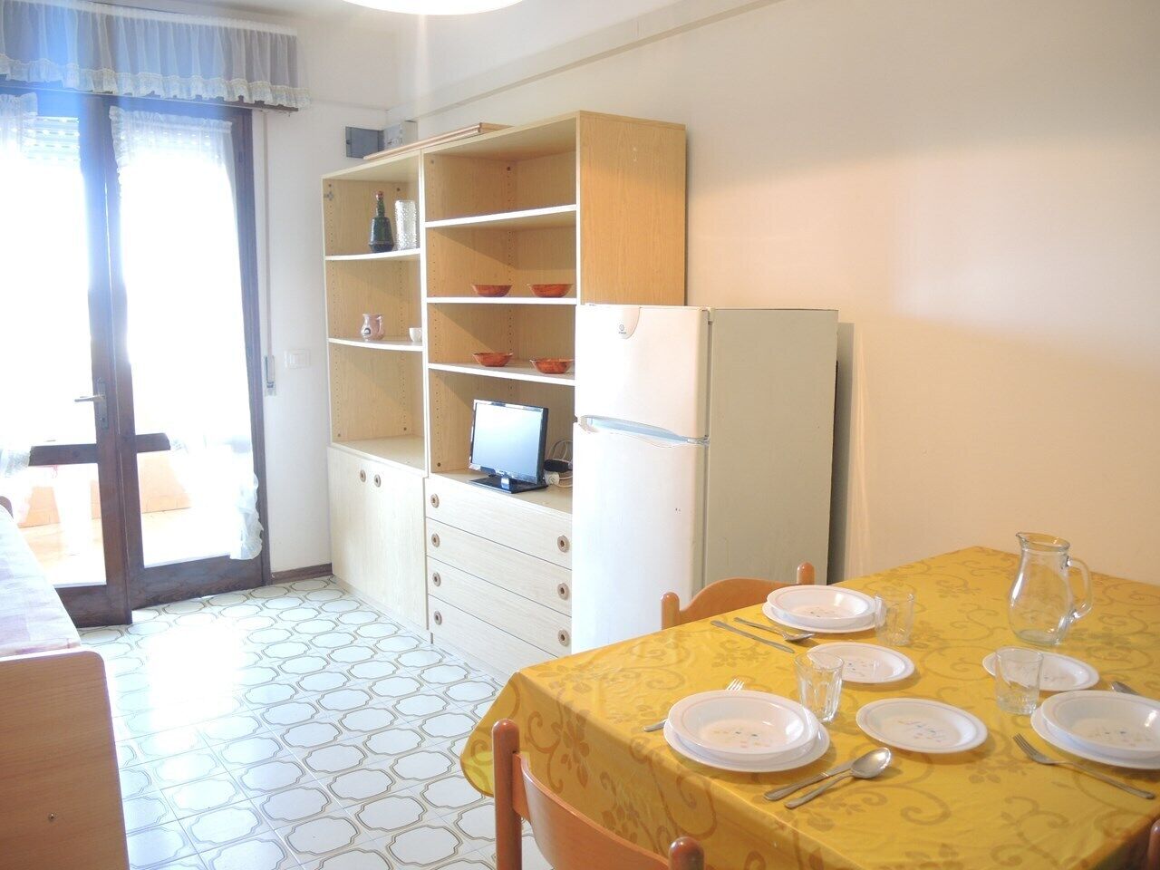 Comedor con una mesa dispuesta para cuatro, un refrigerador y un mueble de madera con estantes.