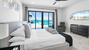A modern hotel room with a large bed, a TV, a ceiling fan, and a view of the beach. - KPOW on the Beach - Brand New 5 bedroom Luxury Beachfront House! (Steventon)