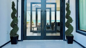 A modern building entrance with a glass door, potted plants, and a view of the sea. - KPOW on the Beach - Brand New 5 bedroom Luxury Beachfront House! (Steventon)