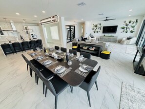 Un comedor moderno con una mesa larga dispuesta para seis personas, una cocina al fondo y una sala de estar con sofá y televisor.