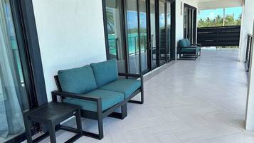 A modern outdoor seating area with a sofa and a small table, large sliding glass doors, and a view of a tropical landscape.