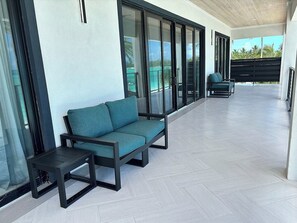 Ein moderner Außenbereich mit einer Couch, einem kleinen Tisch, großen Schiebeglastüren und Blick auf eine tropische Landschaft.