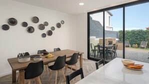 Uma área de refeições com mesa de madeira, cadeiras pretas e toalha branca. Há pratos e copos sobre a mesa. O ambiente possui uma janela grande e vista para o exterior.