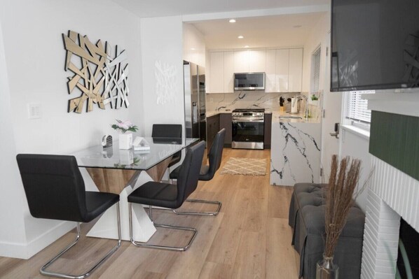 A modern kitchen with a built-in oven and microwave, a dining area with a glass table and black chairs, and a living area with a sofa and decorative plants.