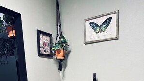 Un petit coin cuisine avec une table en bois, deux tabourets de bar, une plante suspendue et une œuvre d’art représentant un papillon, encadrée et accrochée au mur.